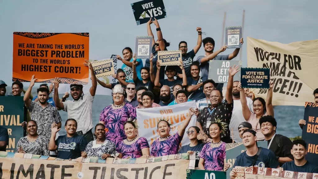 The image shows a large group of mostly young people standing close together, smiling or cheering at the camera. They are holding colorful signs and banners with slogans such as ‘We are taking the world’s biggest problem to the world’s highest court!’, ‘Vote Yes for Climate Justice’ and ‘Our Survival’. Some banners are hand-painted, others printed. In the background, the sky and the sea can be seen. The atmosphere appears energetic and defiant, with the central focus on the demand for climate justice.