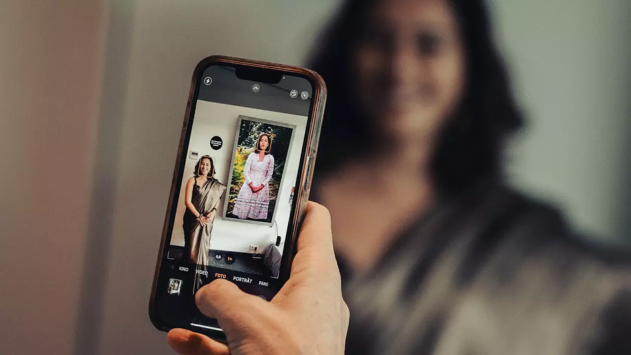 The image shows a person taking a photo with a smartphone. On the phone’s screen, a woman in a sari is standing next to a digital installation displaying another woman in a long dress. In the background, the photographed woman can be seen blurred.