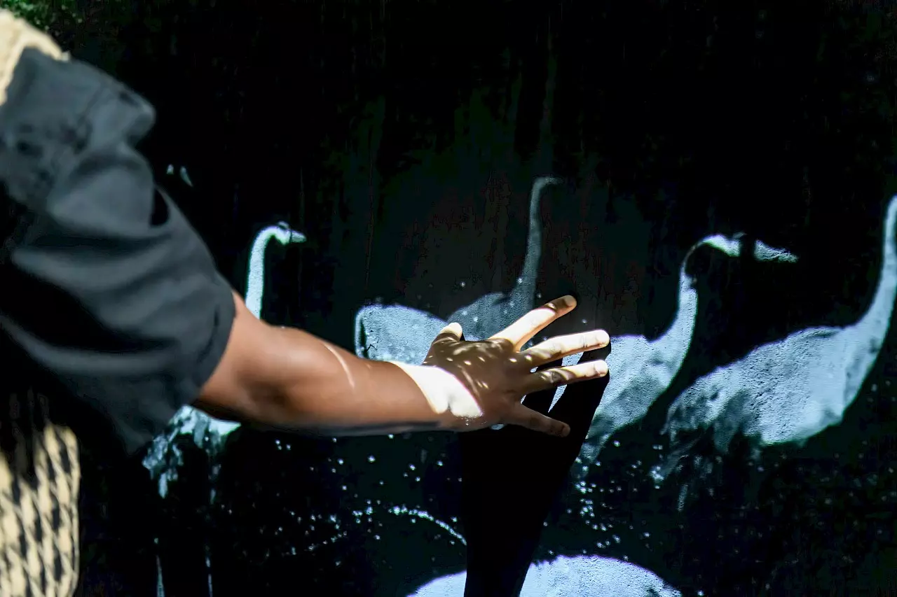 The image shows the outstretched arm of a person whose hand touches a dark projection surface. On the surface, illuminated silhouettes of birds resembling cranes can be seen. Light reflections and shadows play across the hand, creating an impression of interaction between the body and the projection.
