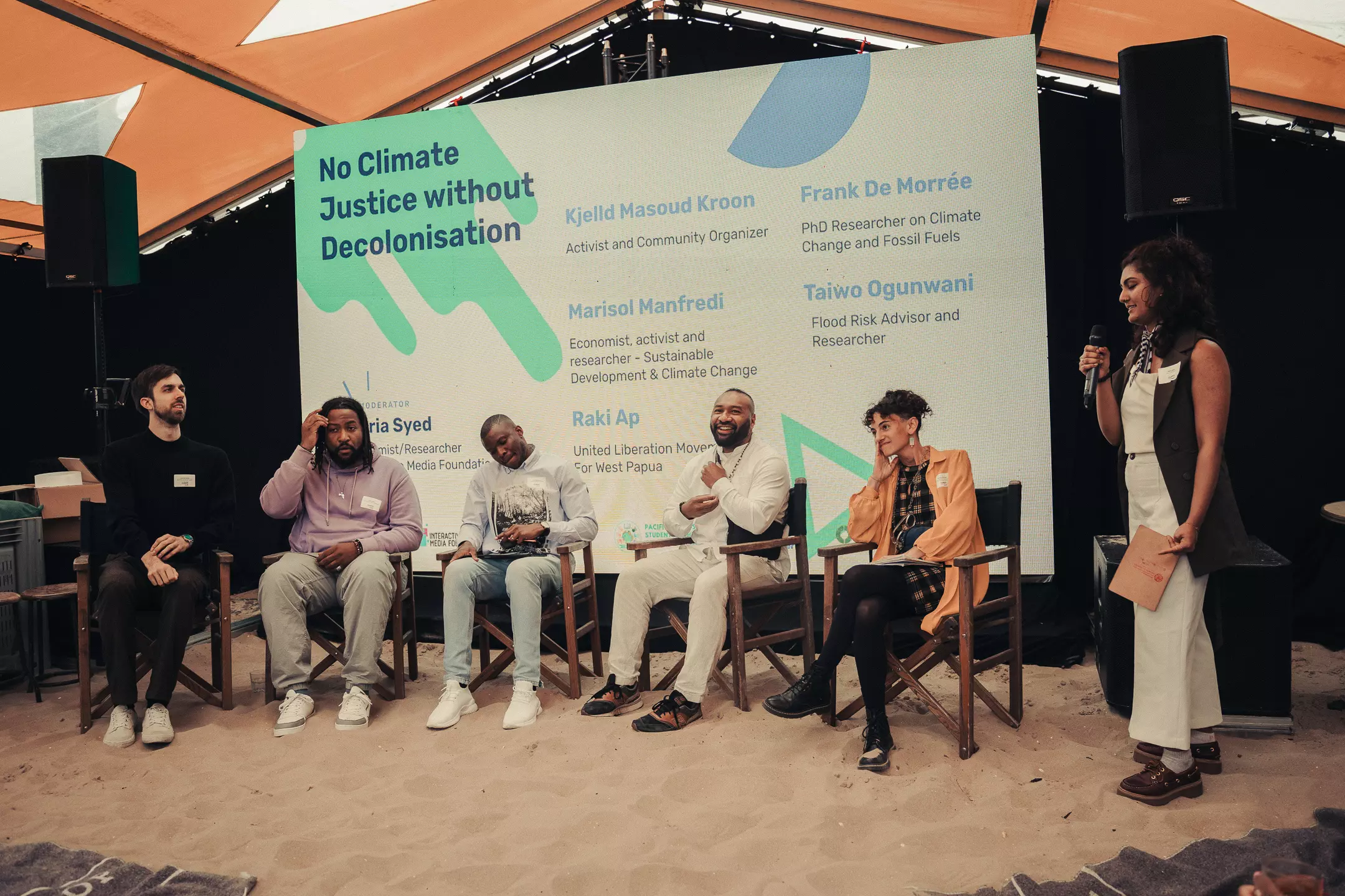 The image shows a panel discussion with six people on stage. In the background, a large screen displays the title “No Climate Justice without Decolonisation.” The screen lists the names and roles of the panelists, including activists, researchers, and advisors from different fields. The moderator stands on the right holding a microphone and speaking, while the panelists sit in chairs, some smiling, others looking thoughtful. The floor is covered with sand, creating an informal and open atmosphere for the discussion.