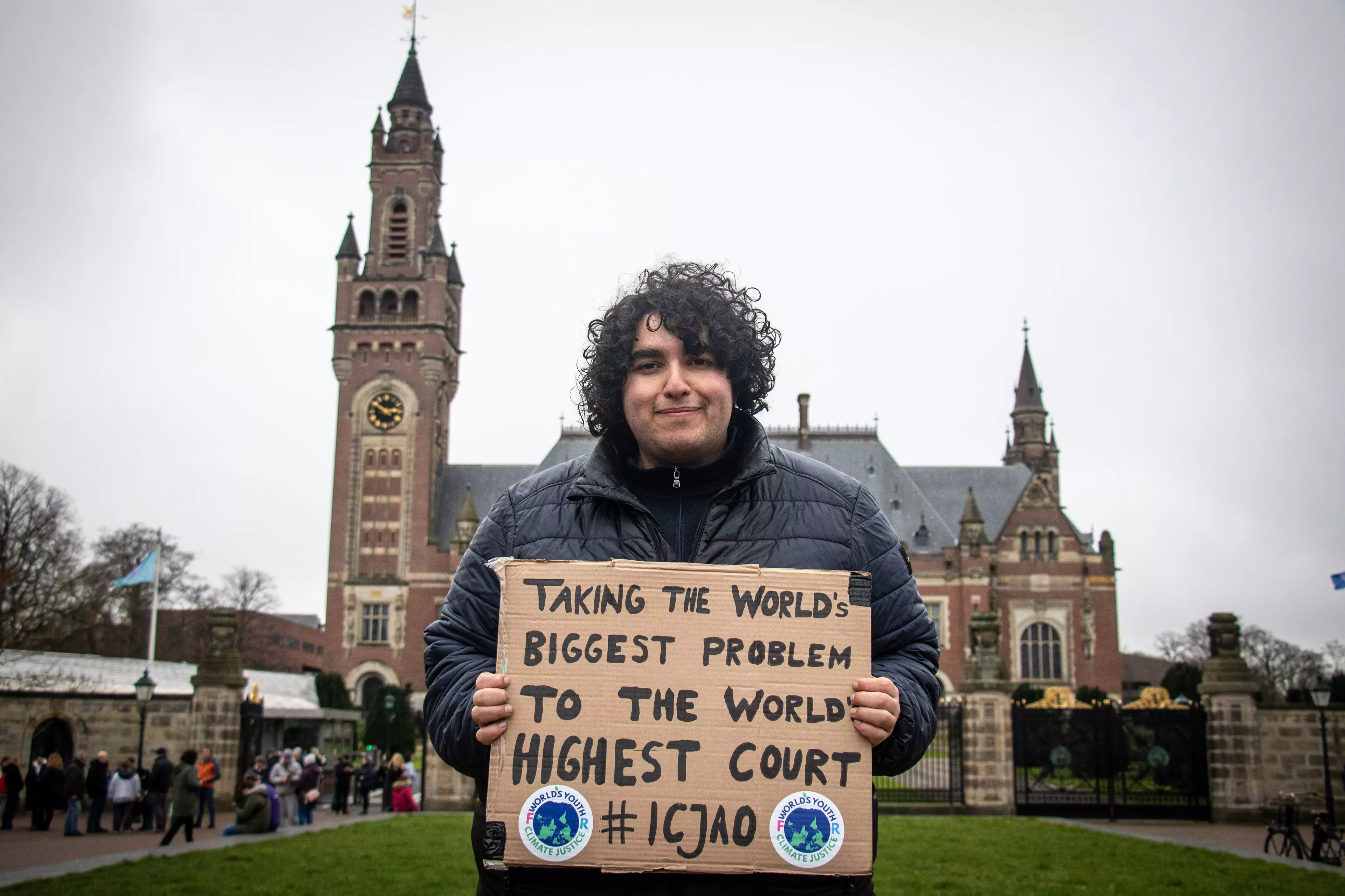 The image shows a young man with curly hair and a dark jacket standing in front of the International Court of Justice in The Hague. He is holding a handwritten cardboard sign that reads “Taking the world’s biggest problem to the world’s highest court #ICJAO.”
