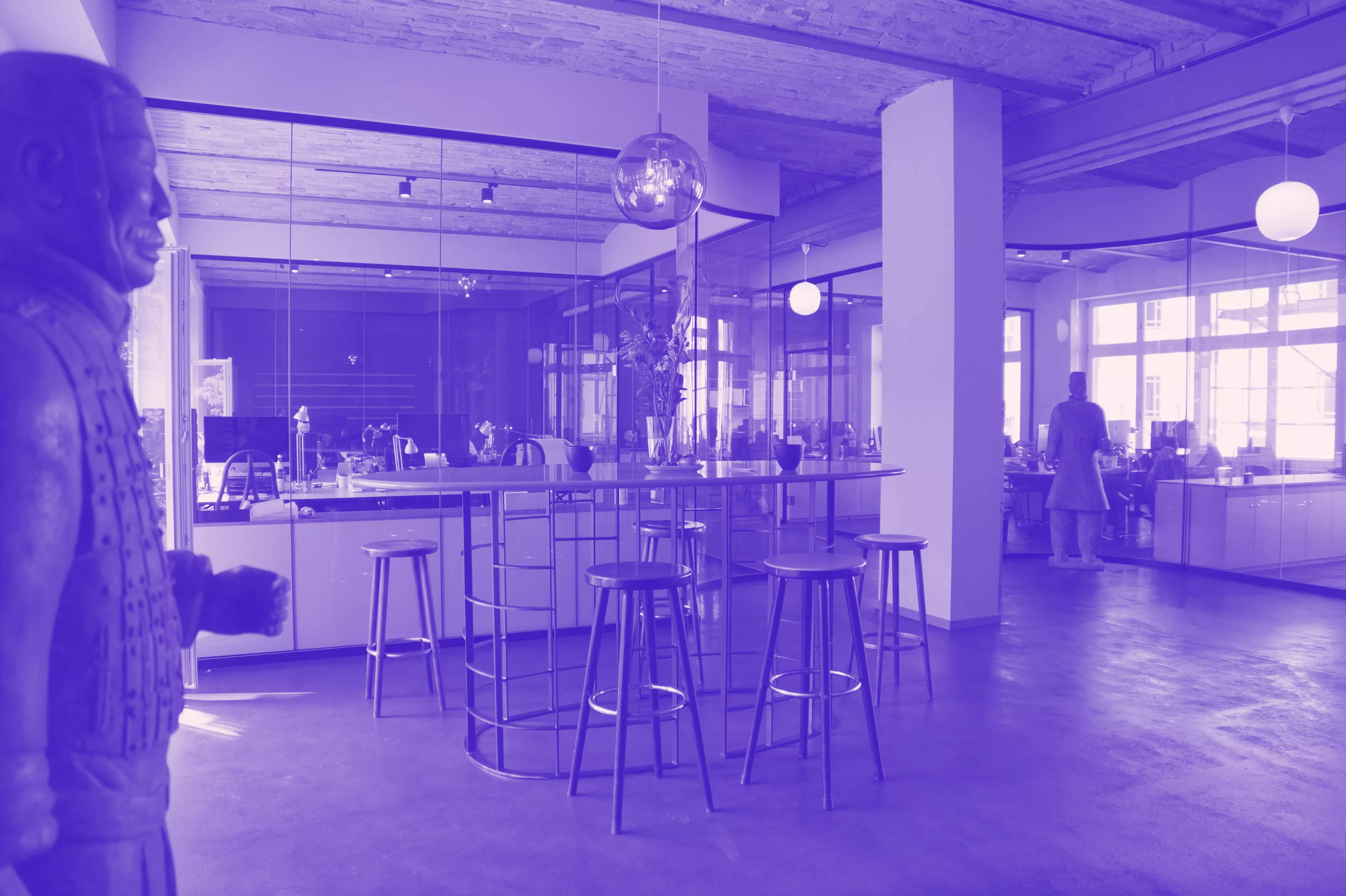 Blick in ein modernes Büro mit Glaswänden, hohen Decken und einem Stehtisch mit Barhockern im Vordergrund. Links und rechts stehen große Statuen, während im Hintergrund Menschen an Schreibtischen arbeiten. Das Bild ist in einem violetten Farbfilter gehalten.