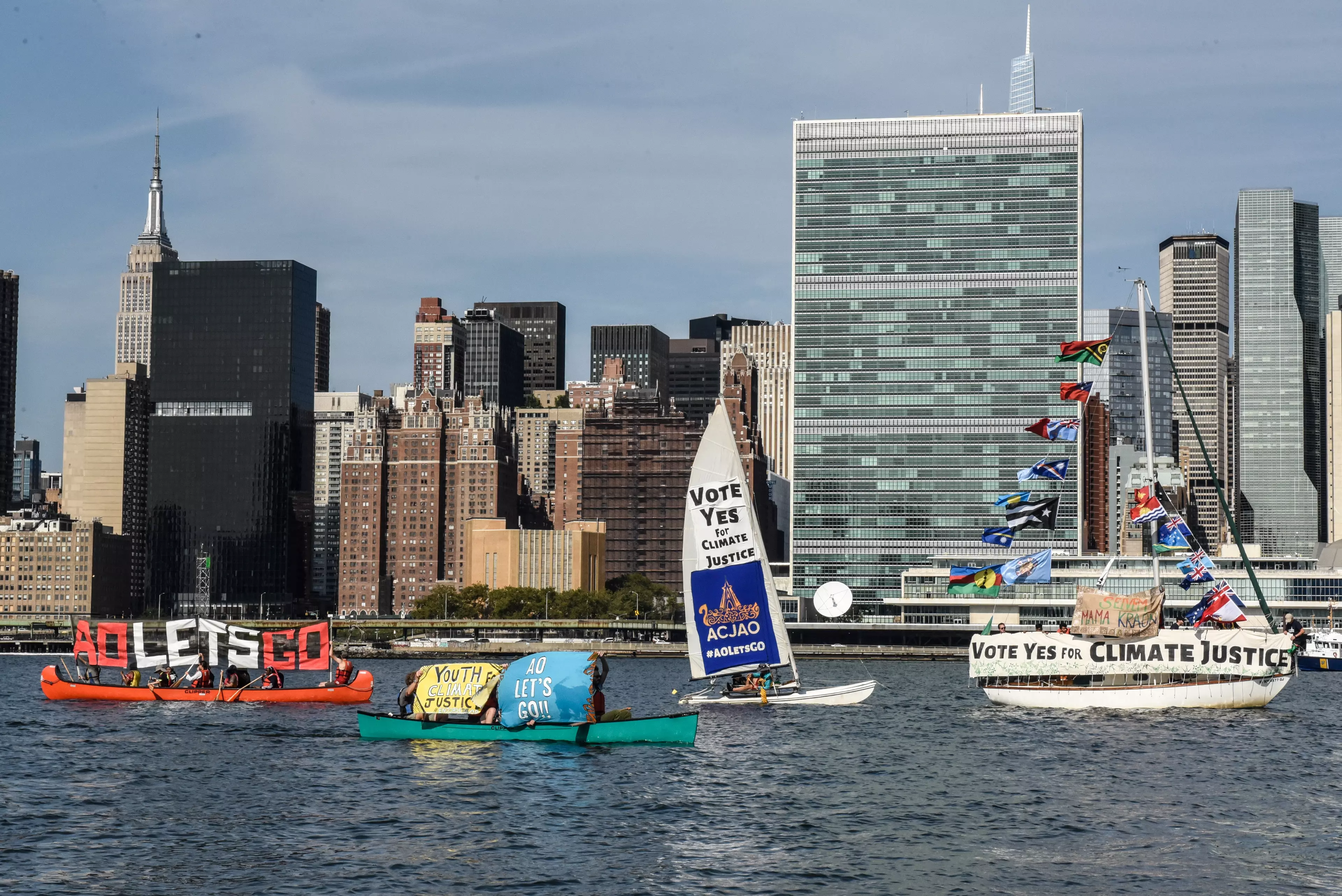 The image shows several boats and canoes on the Hudson River in New York, carrying banners and sails with messages such as “VOTE YES FOR CLIMATE JUSTICE,” “AO LET’S GO,” and “YOUTH CLIMATE JUSTICE.” In the background, the Manhattan skyline is visible, including the UN Headquarters and the Empire State Building. The flotilla draws attention to the ICJAO campaign for climate justice.