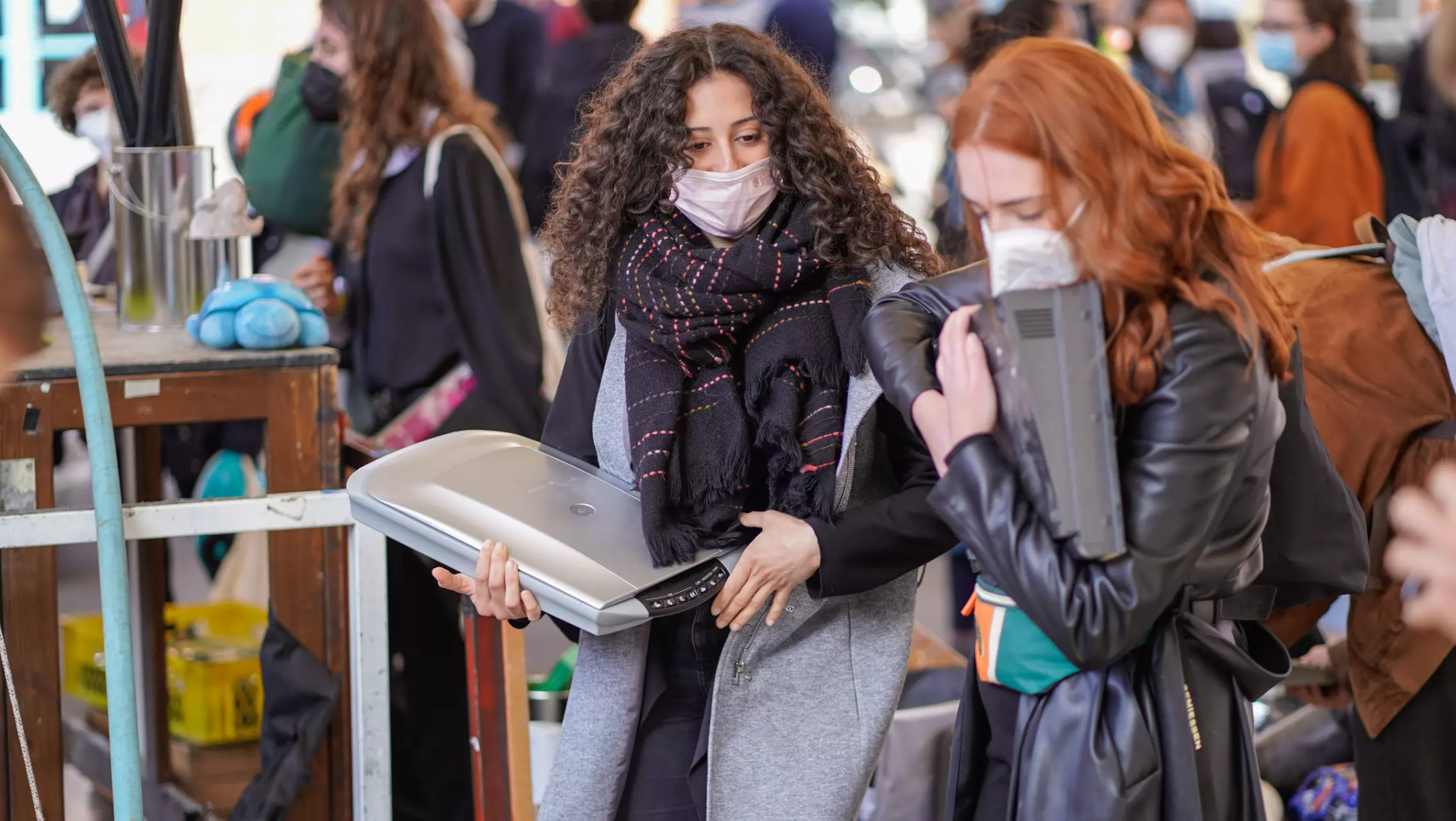 Two young women carry old electronic devices, likely for a repair or recycling project. Both are wearing masks and move through a busy space with other people visible in the background.