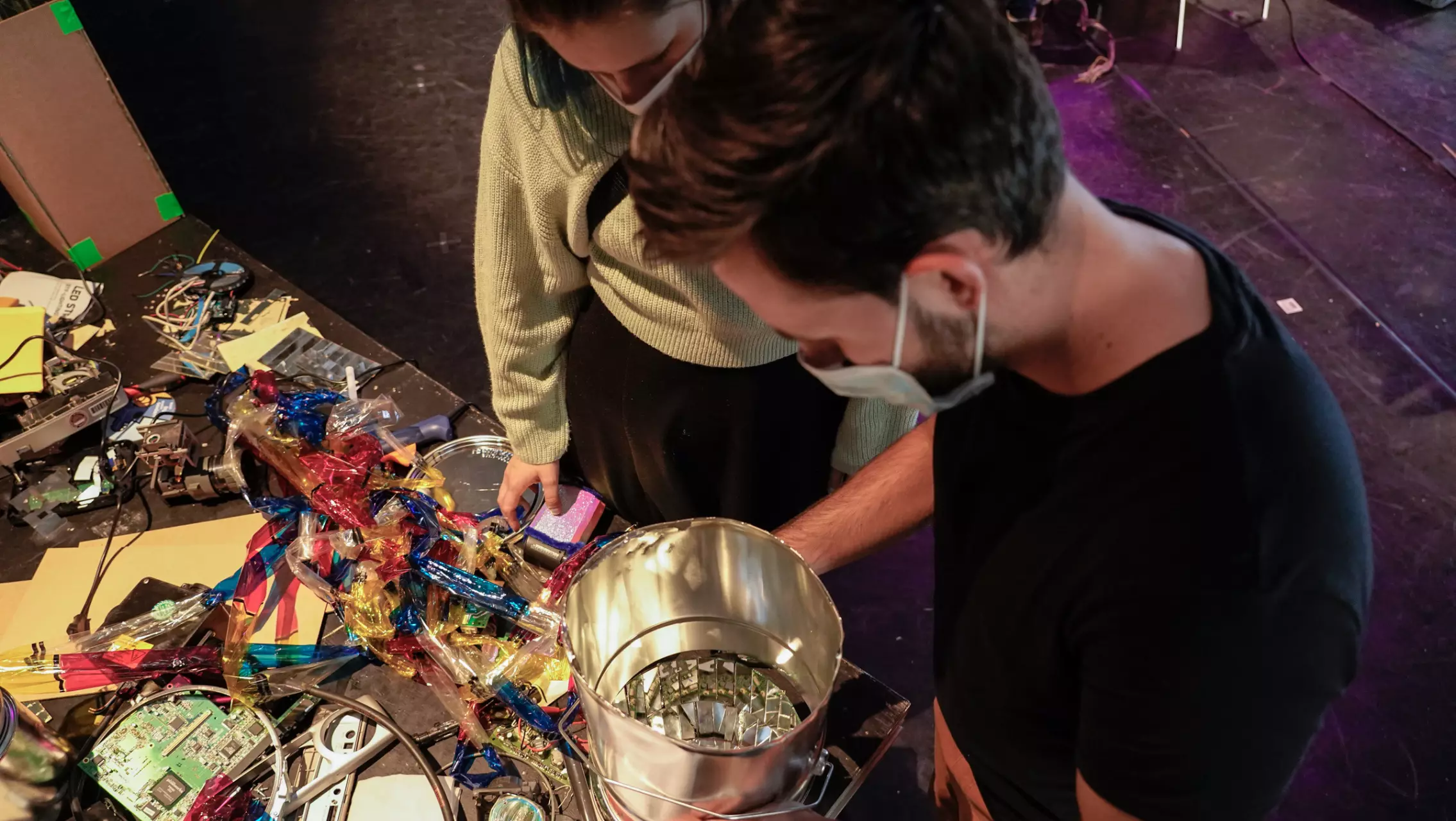 Two young adults wearing masks stand at a crafting table, examining a silver metal bucket that appears to be part of a creative project. The table is covered with colorful foil, wires, circuit boards, and various workshop materials.