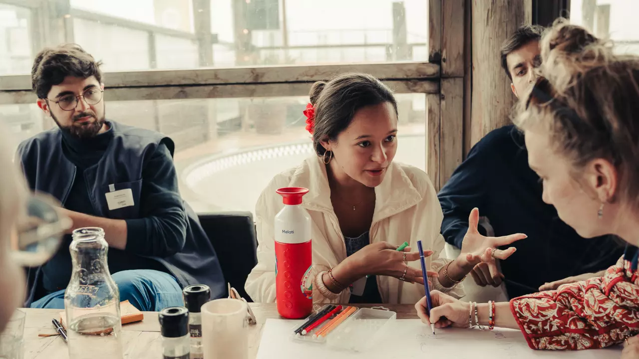 The photo shows a group of four people in a bright room with large windows. At the center sits a young woman wearing a white jacket, speaking with expressive gestures. In front of her are pens, paper, and a water bottle. To the left, a young man with glasses listens attentively, while on the right two other people, partly cropped, are involved in the conversation. The table is scattered with water bottles, cups, and writing materials.