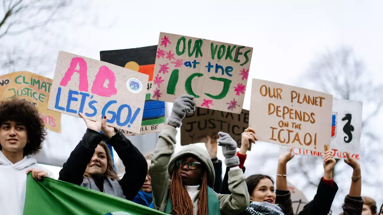 The image shows a group of young people holding up signs at a demonstration. The posters display slogans such as “AO LET’S GO!”, “OUR VOTES at the ICJ,” and “OUR PLANET DEPENDS ON THE ICJAO!”. The protesters are demanding climate justice and highlighting the importance of the International Court of Justice (ICJ) and the ICJAO campaign.