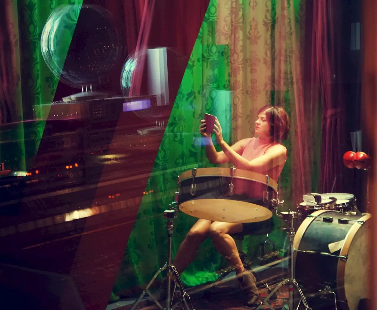 A woman is seated at a drum set in front of colorful green and red curtains. She is holding a smartphone or small device, looking focused. Reflections in the glass layer the image with lights, keyboards, and technical equipment, creating a surreal, multi-layered atmosphere.