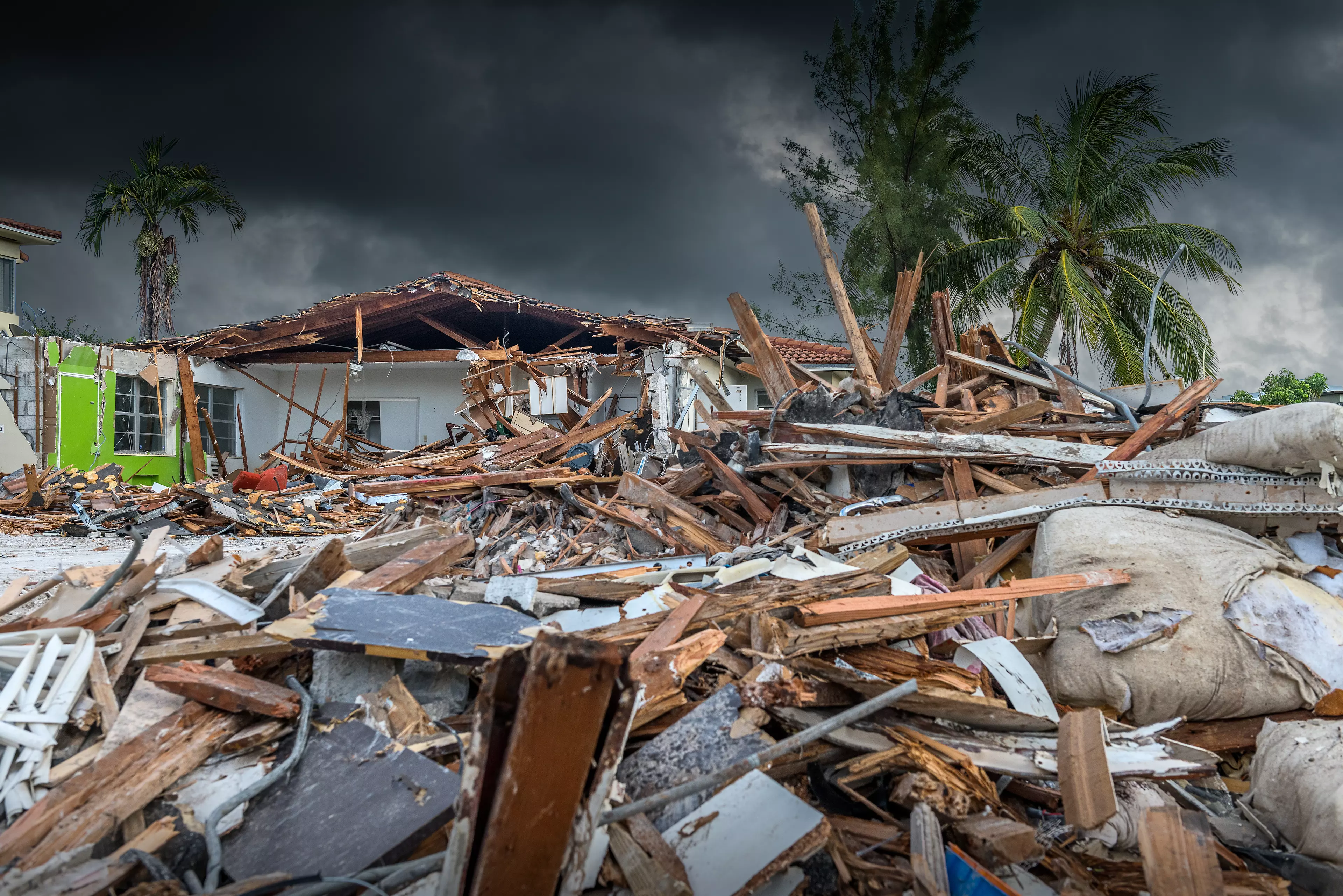 The image shows a heavily damaged house with its roof partially collapsed. In the foreground, large amounts of wood, rubble, and debris are scattered. Palm trees stand in the background, while a dark, threatening sky looms overhead. The scene conveys devastation, likely caused by a storm, hurricane, or other extreme weather event.