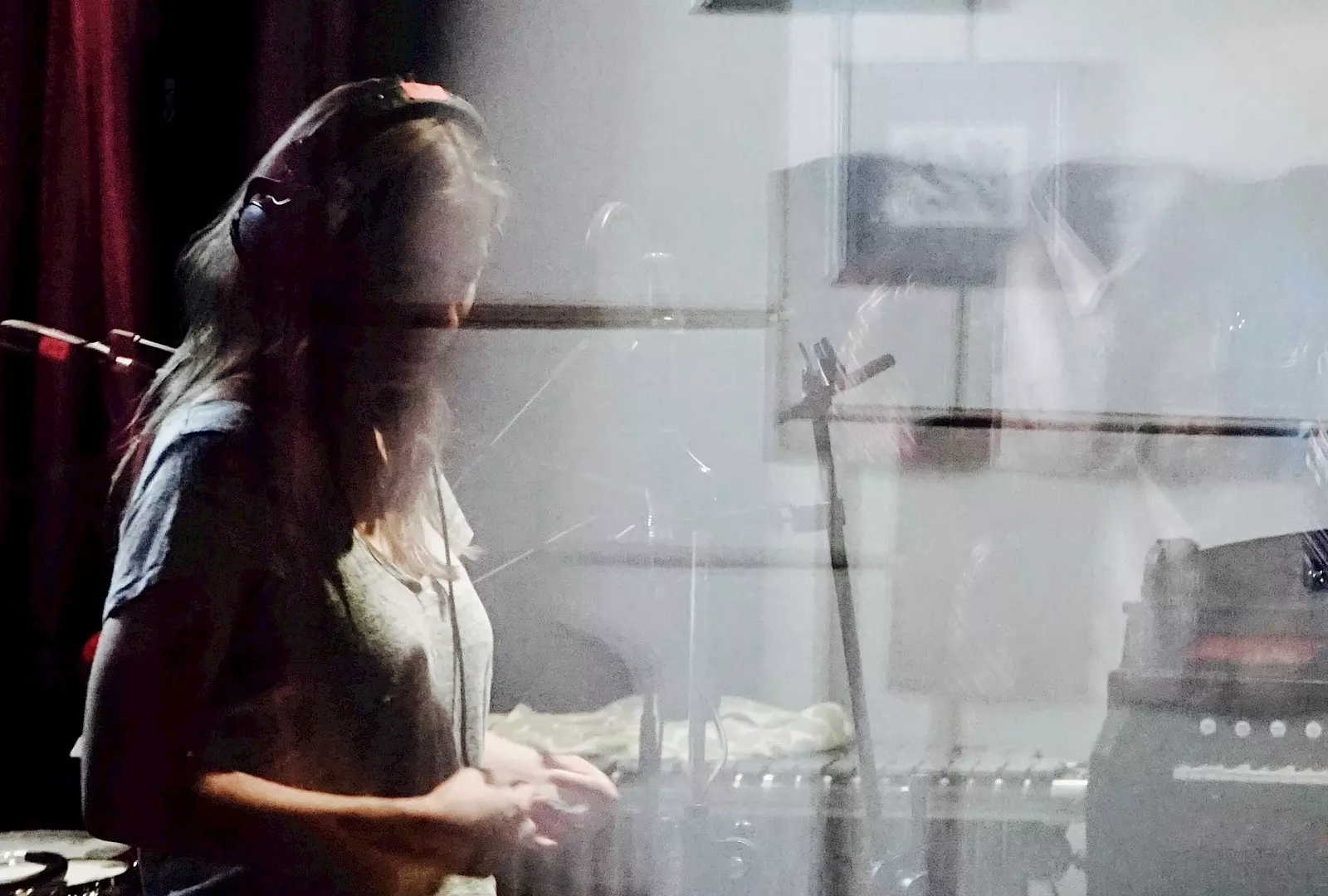 A person wearing headphones stands in a recording studio, surrounded by microphones and instruments. Reflections and overlays on the glass create a dreamy, fragmented atmosphere.