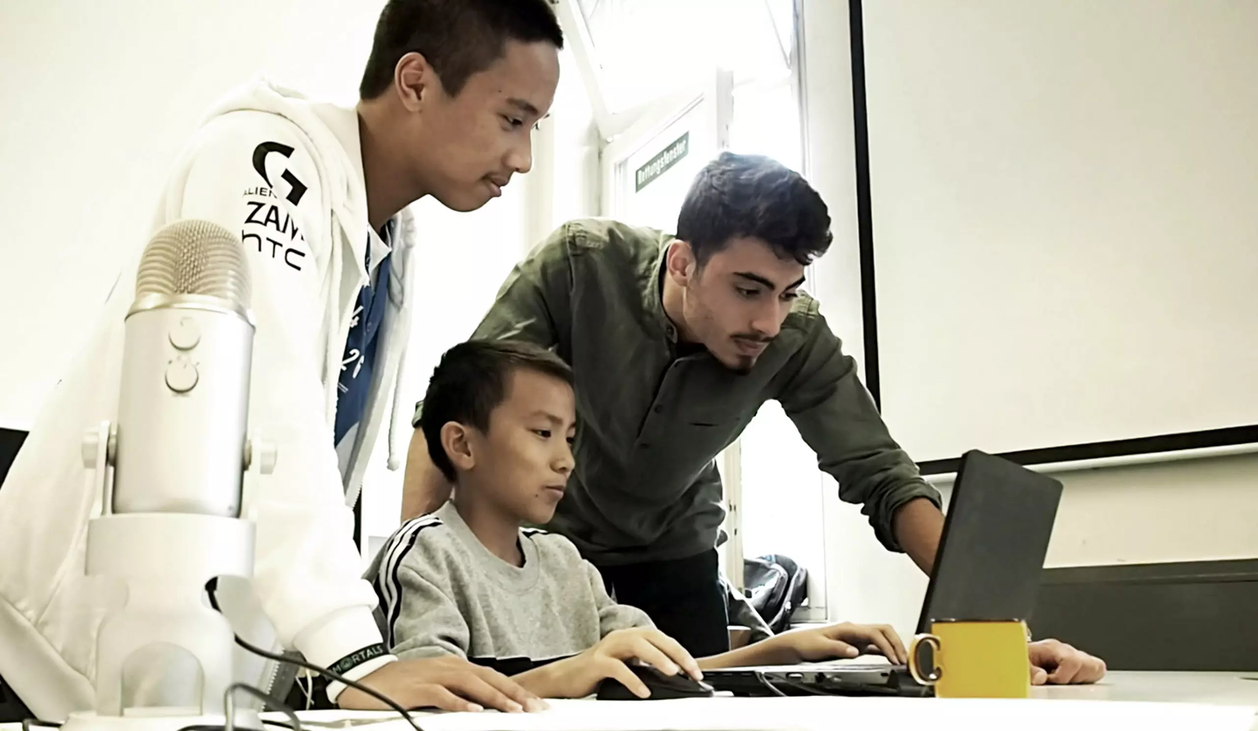 Three young people are working together at a laptop. One is seated, focused on the screen, using the mouse and keyboard, while the other two stand next to him, watching attentively. On the table sits a large microphone and a yellow mug.