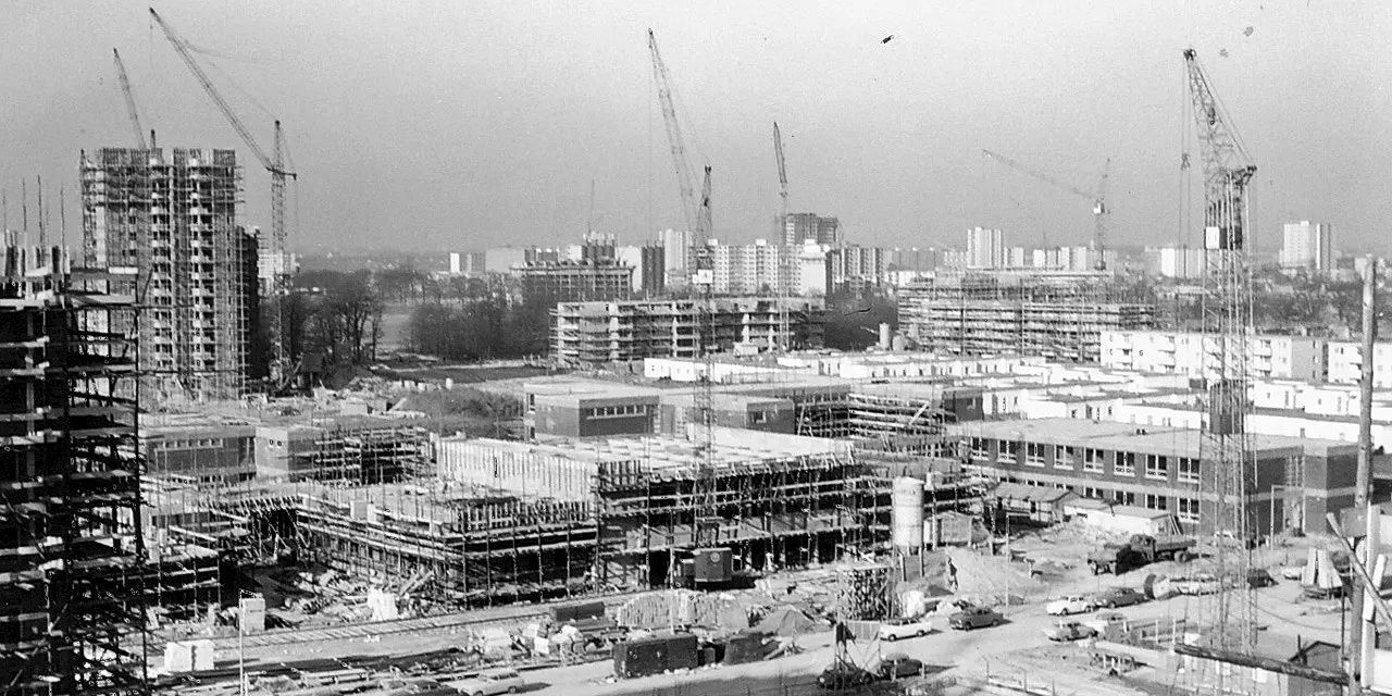 Black-and-white photograph of a large construction site in a city. Several cranes tower above, while numerous buildings are still under construction. In the background, completed high-rise buildings can be seen, reflecting the typical post-war architecture.