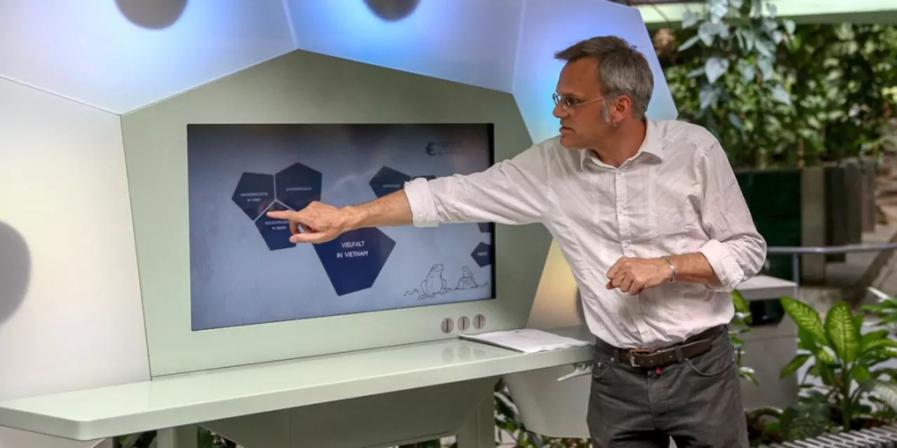 A man in a white shirt explains an interactive exhibition by pointing at a large screen displaying topics on biodiversity in Vietnam.