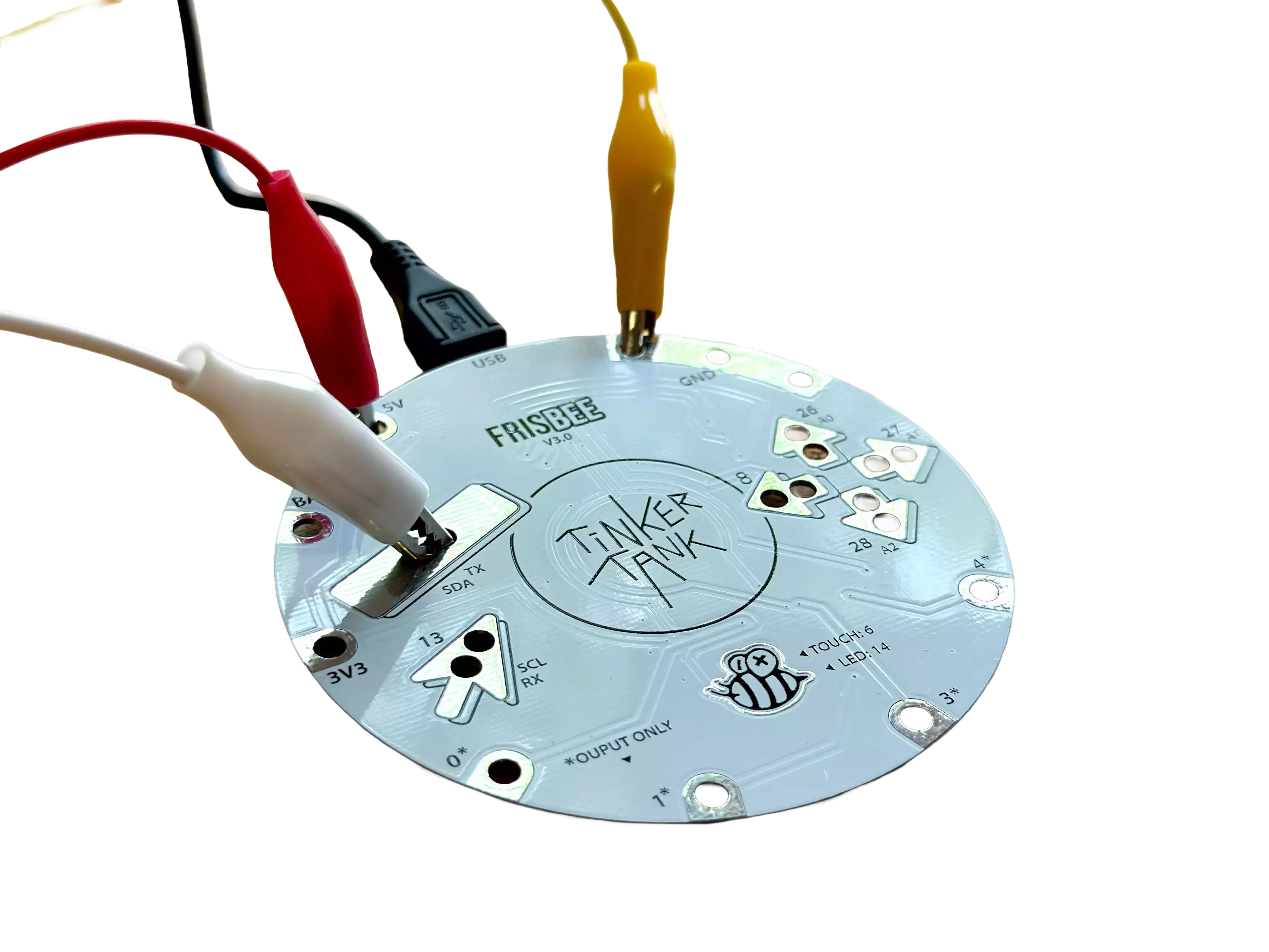 Circular circuit board with the inscription “Tinker Tank” in the center, connected to several cables with alligator clips (white, red, black, yellow). The board is labeled “FRISBEE V3.0” and features various connectors and symbols.
