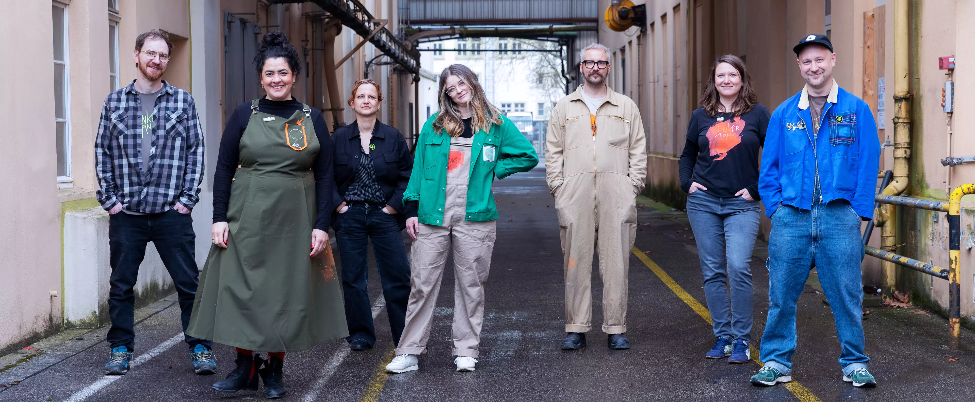 A group of seven people stands in an alleyway between two buildings. They are wearing different outfits – ranging from workwear and overalls to casual shirts and tops – and look relaxed at the camera. The image has the feel of a team portrait that highlights cohesion and diversity.