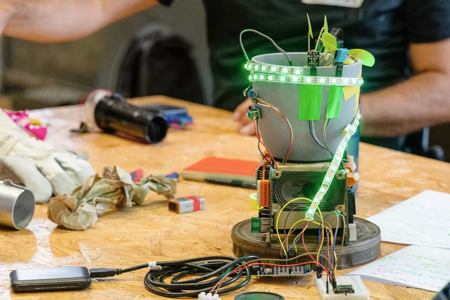 On a wooden table stands a gray flowerpot decorated with green LED light strips, wires, and electronic components. Small plant leaves emerge from the pot, with sensors and circuit boards attached around it. Surrounding the pot are cables, notes, tools, and crafting materials.