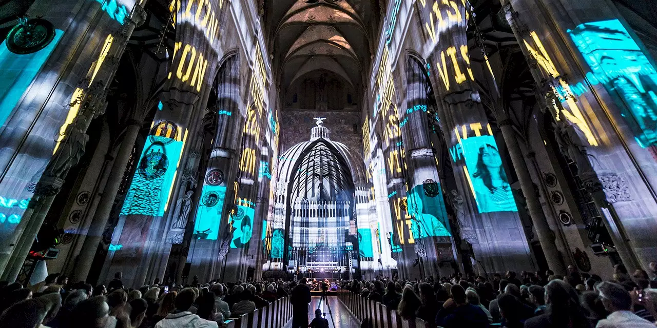 Projections of texts, portraits, and graphic elements illuminate the interior of Ulm Minster during a multimedia performance, in front of a seated audience.