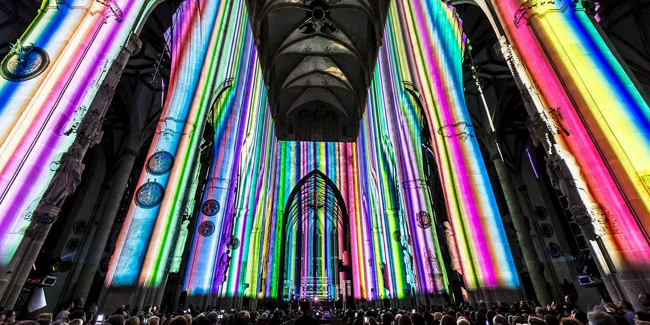 Interior of Ulm Minster during a multimedia show, with colorful vertical rainbow light projections illuminating the tall columns and walls, in front of a large seated audience.