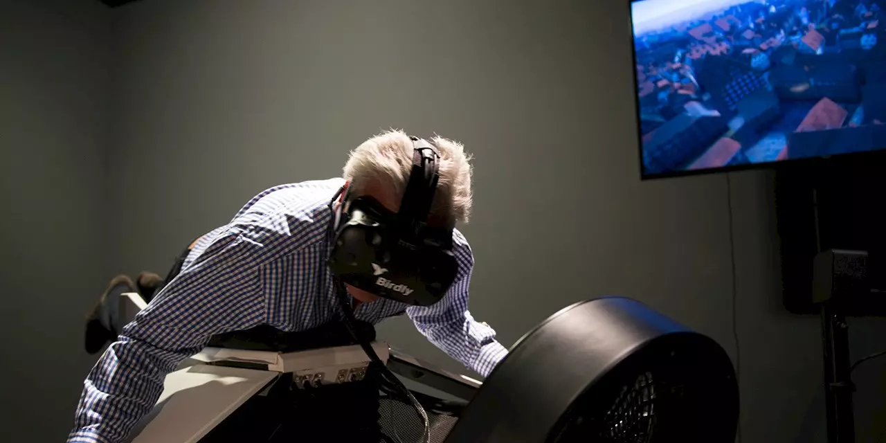 An older man wears a VR headset and lies in a flight simulator designed to simulate the feeling of flying. A screen in the background shows a virtual city view with red rooftops.